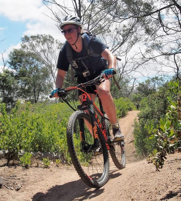 A female mountain biker riding down hill, wearing a dark blue top on a black and orange bike.
